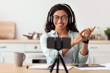 Öğretmen hocası, video konferansı, online sohbet, uzaktan kumanda. Bin yıllık mutlu Afro-Amerikan kadın gözlüklü ve kulaklıklı el kol hareketi yapıyor ve mutfağın içindeki telefon kameralarına bakıyor.