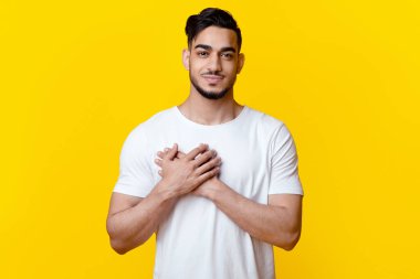 Gülümseyen Arap adam göğsünde avuçlarını tutarak şans, sağlık, aşk ve refah diliyor sarı stüdyo arka planında, dua ediyor ya da yardım istiyor.