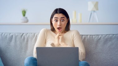 Şok olmuş iş kadını dizüstü bilgisayarın önünde oturmuş ekrana bakıyor. Komik yüz ifadesi duyguları, duygular, algılama sorunları, tepkiler. Evdeki kanepede el kol hareketi yapan genç Arap bayan şaşırdı.