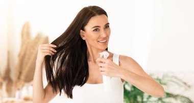 Saç stili, kozmetik ürünler, bölünmüş kuru uçlar, güneş koruması. Mükemmel ciltli, genç ve güzel esmer kadın beyaz banyo arka planında saç spreyi, termal su, boş alan.