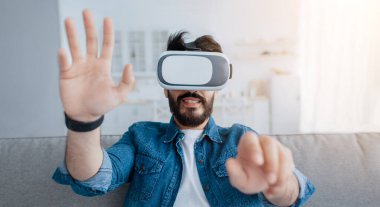 Gelecekteki teknoloji konsepti. Video oyunları oynayan, sanal gerçeklik gözlüğü takan, el kol hareketi yapan, evdeki kanepede oturan bir adam. Genç adam modern eğlence ekipmanları kullanıyor, hafta sonları eğleniyor.