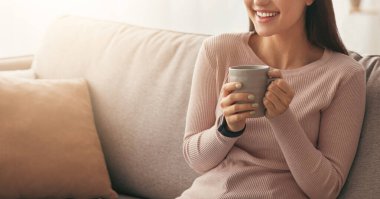 Evde boş zaman geçirmek, rahatlamak ve kendini izole ederken sıcak içkiyle dinlenmek. Neşeli genç Arap kadın sessizliğin tadını çıkarıyor ve minimalist oturma odasındaki kanepede oturup kahve içiyor.