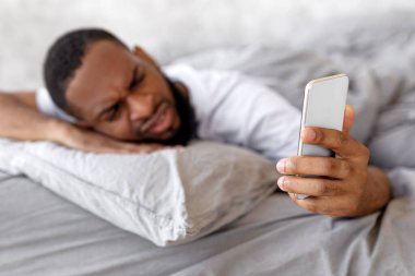 Bir adam rahat bir odada yastığın üzerinde dinleniyor ve hayal kırıklığına uğramış bir ifadeyle akıllı telefonuna bakıyor. Teknolojinin zorluklarını yansıtan rahat bir an..