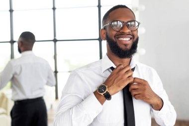 Beyaz gömlekli ve siyah kravatlı bir adam modern, aydınlık bir odada kravatını düzeltirken gülümsüyor. Arka planda kıyafetlerini ayarlayıp atmosfere ekleyen başka bir birey görülebilir..