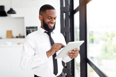 Beyaz gömlekli ve siyah kravatlı bir adam çağdaş bir ofiste büyük bir pencerenin yanında duruyor. Bir tablete odaklanmış, neşeli bir ifade sergiliyor, işiyle ilgili olduğunu gösteriyor..