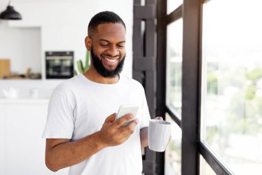 Sakallı bir adam akıllı telefonuna bakıp diğer elinde bir kupa kahve tutarken gülümsüyor. Parlak mutfakta büyük pencereler ve modern dekorlar yer alıyor..