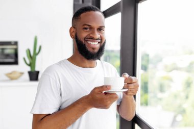 Çağdaş bir mutfakta elinde bir fincan kahveyle büyük bir pencerenin yanında duran ve gülümseyen bir adam. Güneş ışığı odayı dolduruyor, yakındaki bitkilerin yeşilliğini vurguluyor..