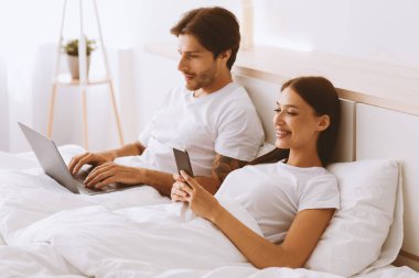 Genç Avrupalı kadının sosyal ağlarda telefonda sohbet etmesi, adamın yatak odasında dizüstü bilgisayarda çalışması. Modern ilişkiler ve çevrimiçi iletişim, boş zamanlardaki aygıt, yatmadan önce sohbet