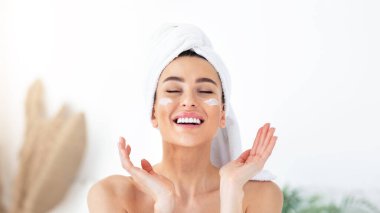 Cilt bakımı, temizlik, eko organik kozmetik, spa, evde rahatlamak ve yeni normal, sosyal mesafeler. Yüzünde krem, beyaz banyo arka planında jestleri olan havluyla gülen bin yıllık tatlı bir kadın.