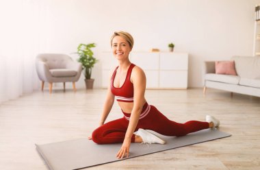 Antrenmandan önce ısınıyorum. Gülümseyen sporcu kadın evde antrenman yapıyor, yoga minderi üzerinde güzel kadın bacakları esnetiyor, mavi sutyen, tayt ve ayakkabı giyiyor, kameraya bakıyor.