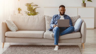 Gülümseyen siyah sakallı adam oturma odasındaki kanepede dizüstü bilgisayarda çalışıyor. Mesafe işi, serbest çalışma, sosyal ağlarda iletişim ve evdeki teknolojiyle çevrimiçi oyunlar