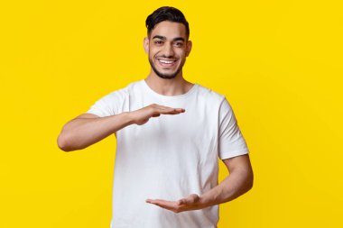 Positive young muslim guy in white t-shirt and jeans holding something invisible in his hands next to his abdomen and smiling at camera, posing on yellow studio background, copy space