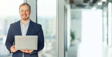 Takım elbiseli gülen adam dizüstü bilgisayar tutuyor ve kameraya gülümsüyor, modern ofis arka planı, fotokopi alanı olan bir panorama.