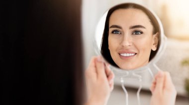 Yaşlanma önleyici, Cilt bakımı, kovid-19 kilitlenmesi sırasında evde. Kozmetoloji, güzellik terapisi ve güvenlik. Gülümseyen milenyum dişisi küçük aynaya bakar ve modern yatak odasının içinde güzel bir yüzün tadını çıkarır.