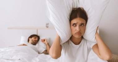 Yoksul Avrupalı kadın, yatak odasındaki yatak odasındaki kocanın horlamasından muzdarip, yastıkla kulaklarını örtüyor. Geceleri dayanılmaz horlama ve gürültü, uykusuzluk ve apne.