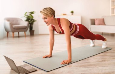 Mavi Giyinen Gülümseyen Kadın Spor Giysileri Egzersizi Yaparken Tam Vücut Uzunluğu Ya da Oturma odasında Yoga Mat Üzerinde Kalas Konumu Kişisel Eğitmenle Video Görüşmesi Yaparken