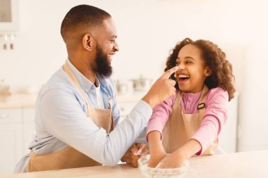 Kara Baba ve küçük çocuğu mutfakta birlikte yemek pişirirken eğleniyorlar. Mutlu Afro-Amerikan ailesi evde yemek pişirirken birbirleriyle oynaşıyor, Baba Kızların Burnuna dokunuyor