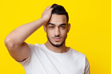Çekici, sakallı, beyaz tişörtlü ortadoğu çocuğu sarı stüdyo arka planında poz verirken saçına dokunuyor, bayanlarla flört ediyor, yakın plan portresi, fotokopi uzayı kopyalıyor.