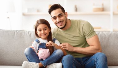 Kişisel Tasarruf ve Bütçe Planlama. Yakışıklı Arap baba ve kızı kumbaraya para koyuyorlar evdeki koltukta oturup kameraya bakarak yatırım yapıyorlar. Çocuk ve Mali Edebiyat