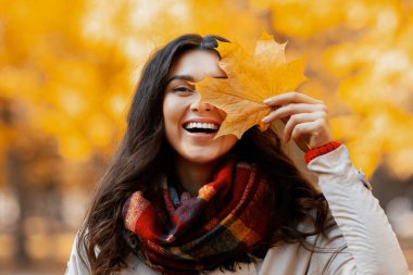 Güzel bir milenyum kadını, sonbaharda parkta yürürken gülerek ve sarı sonbahar yaprağıyla gözlerini kapayarak. Güzel güneşli bir günde dışarıda vakit geçiren mutlu genç bayanın portresi.