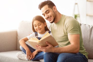 Yetiştirilme ve ebeveynlik kavramı. Evde kitap okuyan genç baba ve kız, bedava fotokopi alanı. Yakışıklı baba ve çocuk hafta sonunun tadını çıkarıyor oturma odasında rahat bir kanepede oturuyor.