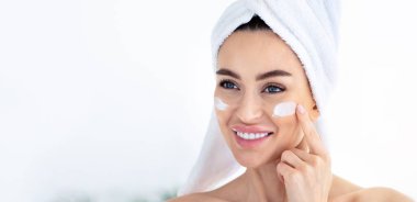 Güzel lüks kozmetik prosedürler, spa ve kozmetik. Havlu tutan mutlu milenyum kadını beyaz banyo arka planı, panorama, fotokopi alanı, yüz yüze krem sürer ve uygular.