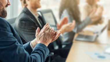 Bir grup iş adamı bir konferans masasının etrafında oturmuş takdirlerini alkışlıyorlar. Masada bir dizüstü bilgisayar ve kağıtlar görülüyor.