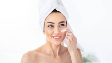 Güzel genç bir kadın taze deri için maske yapıyor. Kadın güzellik yüz bakımı, yüz bakımı. Mutlu milenyum havlusu olan güzel bayan nemlendirici krem sürüyor, beyaz banyo arka planında, boşluğu kopyala