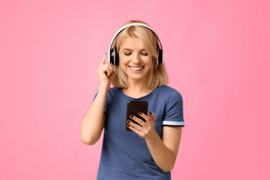 En sevdiğiniz eğlence. Kablosuz kulaklıklı mutlu bir kadın akıllı telefondan müzik dinliyor, bej arka planda yeni uygulamanın tadını çıkarıyor, stüdyo çekimi.