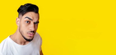 Top view of surprised handsome bearded young arabic guy in white t-shirt looking at camera with eyebrow up, posing on yellow studio background, closeup photo, copy space for advertisement