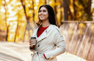 Zarif kıyafetli bin yıllık kadın sonbahar parkında yürüyüş yaparken kahvesini yudumluyor. Gülümseyen genç bayan, güneşli bir sonbahar gününde açık havada sıcak içecek içiyor.