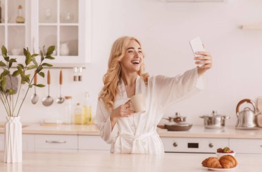 Cüppeli, güzel, Avrupalı, genç, sarışın bir kadın kahve içiyor, kahvaltıda, İskandinav mutfağının iç kısımlarında akıllı telefondan selfie çekiyor, fotokopi çekiyor. Modern aygıt, uygulama, blog ve sosyal ağlar