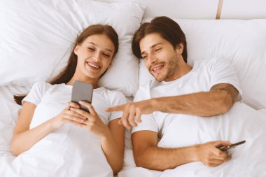 Neşeli Avrupalı genç bir aile telefona bakıyor, sosyal ağlarda sohbet ediyor, yatak odasının içinde yatakta sörf yapıyor. Birlikte komik videolar izlemek, covid-19 salgını sırasında online alışveriş yapmak.