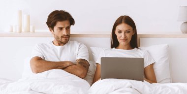 Huysuz, üzgün, gücenmiş, beyaz bir koca kollarını kavuşturmuş, karısına bakıyor, beyaz yatak odasının iç kısmında dizüstü bilgisayarda yazıyor. Covid-19 salgını, kıskançlık, evde çalışma, sosyal ağlar, boş alan