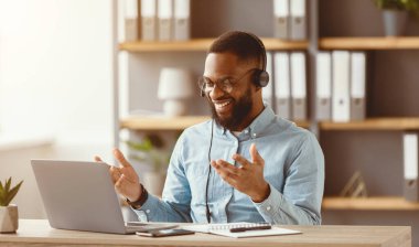 Gözlüklü, kulaklıklı, gülümseyen siyah sakallı erkek. El kol hareketleri, iletişim ve ofis içi dizüstü bilgisayarda çalışıyor. Covid-19 karantina, çalışma, seminer, özel ders ve başarılı iş