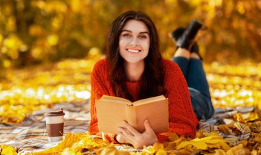 Elinde kitap ve kahveyle gülen genç bir kadın düşen sonbahar yaprakları arasında battaniyeye uzanıp sıcak sonbahar gününün tadını çıkarıyor. Samimi kazaklı mutlu bayan dışarıda heyecanlı hikayeler okuyor.