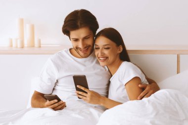 Kafkasyalı mutlu karı koca akıllı telefonlara bakıyor, sarılıyor, yatak odasında birlikte video izliyorlar. Yeni uygulama, harika bir teklif, cihaz bağımlılığı ve covid-19 salgını sırasında sosyal medya