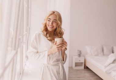 Pozitif sabah, hafta sonunun ve tatilin tadını çıkar, boş zaman. Binlerce yıllık neşeli Avrupalı kadın kahve içiyor ve pencereden dışarı bakıyor. Minimalist beyaz yatak odasında.