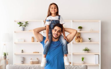 Bağlanma kavramı. Sevimli küçük kızını omuzlarında taşıyan neşeli Orta Doğulu bir adamın portresi, mutlu aile pozu veren, kameraya bakan, oturma odasında dikilirken eğlenen...