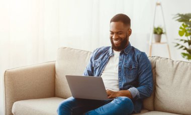 Gülümseyen bin yıllık Afro-Amerikan sakallı adam dizüstü bilgisayarda yazıyor, online video izliyor, oturma odasında internette çalışıyor ve sörf yapıyor. Evde iş, sohbet, telefon, toplantı ve sosyal mesafe