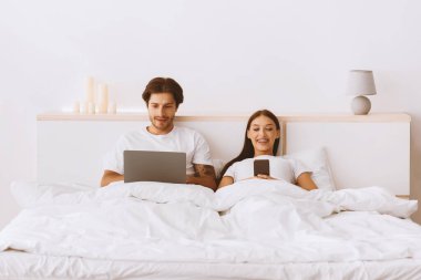 Neşeli Avrupalı genç bir kadın telefonda yazıyor, kocası internette internette internette internette sörf yapıyor beyaz yatak odasının içinde yatağında. Boş zamanlarında sosyal ağlarda sohbet eden çiftler, yeni normal, modern ilişkiler.