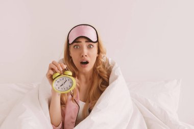 Komik korkmuş bin yıllık beyaz kadın pembe pijamalı ve uyku maskeli, ağzı açık, battaniyeli beyaz arka planda çalar saati gösteriyor. Uyuyakalmışım, işe geç kalmışım ya da toplantıdayım.