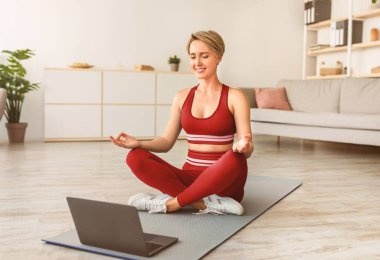 Bilinç, Denge ve Barış Konsepti. Gülümseyen kadın oturma odasında yoga minderinin üzerine oturmuş, parmaklarını Gyan Mudra el hareketleriyle odaklanıyor, bilgisayar kullanarak, sanal ders kullanarak,