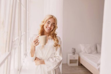Hafta sonu boş zamanlarınızda ya da rahat, minimalist tavan arasında harika bir sabah geçirin. Gülümseyen genç, güzel sarışın kadın uyandığında bir fincan kahveyle, panoramik pencerenin yanında, güneş parlıyor.