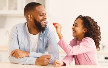 Sağlıklı yemek. Şirin Afrikalı Amerikalı kız, mutfakta taze salata pişirirken mutlu babasını domatesle besliyor. Bağlanma, Sevgi, Babalık ve Seven Aile Kavramı