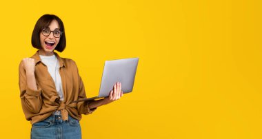 Aşırı mutlu bir kadın yumruğunu sıktı ve sarı stüdyo arka planında duran dizüstü bilgisayarla başarıyı kutladı. Heyecanlı bayan öğrenci zafer ve şans için tezahürat yapıyor.