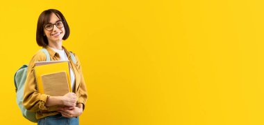 Akademik eğitim. Sırt çantalı ve defterli genç bayan sarı stüdyo arka planında kameraya gülümsüyor. Üniversiteye başlamaya hazır bayan üniversite öğrencisi