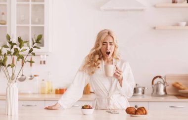 Uykulu, yorgun, genç, beyaz tenli, bornozlu güzel, sarışın bayan erken uyanıyor, kahve içiyor ve modern mutfak içinde esniyor, fotokopi çekiyor. Uykusuzluk, yorgunluk, kötü gece rüyası ve kahvaltı