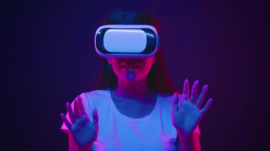 Neon ışıklarının aydınlattığı canlı bir ortamda genç bir kadın sanal gerçeklik kulaklığıyla etkileşime girer. Ellerini kaldırdı, ona yakınlık ve merakını gösteriyor..