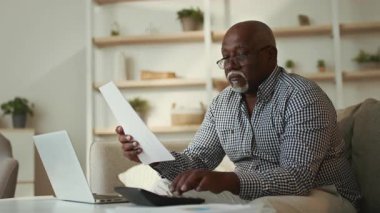 Yaşlı bir Afrikalı Amerikalı adam evinde huzurlu bir öğleden sonra önemli belgeleri incelerken dizüstü bilgisayarına rahatça odaklanarak oturma odasında oturuyor..